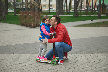 快乐的父亲拥抱他可爱的小女孩-心爱的女儿，亲吻她的脸颊，同时在城市公园散步，在美丽的早春享受骑滑板车