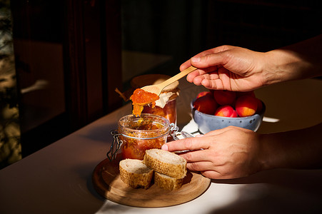 一位家庭主妇的手拿着木勺，在面包上涂抹自制杏酱的特写。
