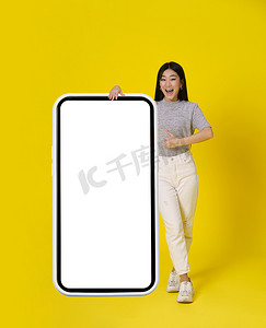 迷人的亚洲女孩竖起大拇指，站在巨大的智能手机旁边，智能手机上有白色屏幕，穿着黄色背景的休闲装。