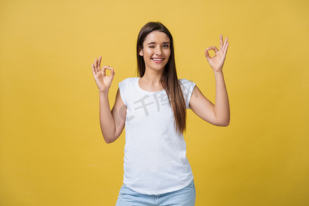 黄色手指手势摄影照片_快乐的年轻女人用手指表现出好的手势，在黄色背景中眨眼