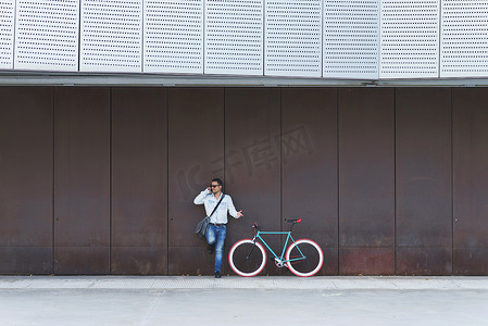 固定的空间摄影照片_戴着太阳镜、背着肩包的年轻人一边打电话一边站在固定齿轮自行车旁