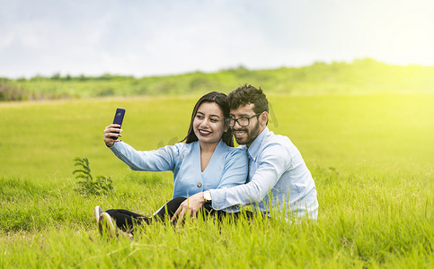 微笑相爱的情侣坐在草地上自拍，相爱的年轻情侣在田野里自拍，相爱的人用智能手机在田野里自拍