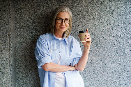 折叠纸杯摄影照片_退休时迷人的成熟灰发女性用纸杯喝咖啡，站在户外，靠在大理石、花岗岩墙上，微笑着，一只手臂折叠着，看着相机