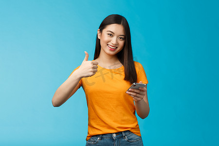 满意可爱的亚洲黑发女郎拿着智能手机竖起大拇指很高兴，推荐很棒的小工具，批准好的新应用程序，同意朋友发布好的博客文章，站在蓝色背景