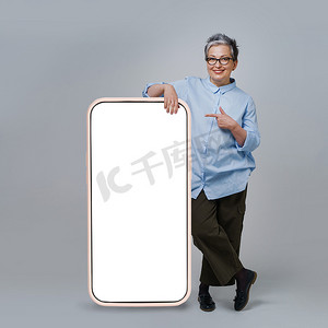 成熟的灰发女人指着巨大的、巨大的智能手机屏幕，白色、空白的屏幕站立，靠在白色背景上孤立的休闲装上。