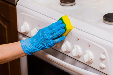戴着家用手套的女家庭主妇的手用海绵擦拭燃气灶控制面板。