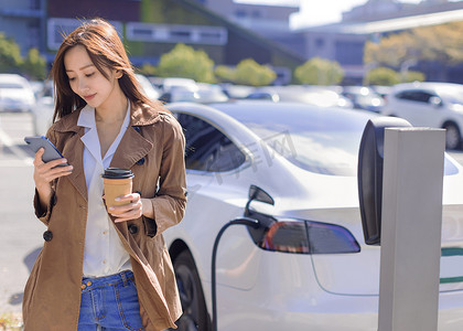 微笑的年轻女性站在电动车附近的城市停车场，从小城市站为汽车电池充电，喝咖啡和使用智能手机