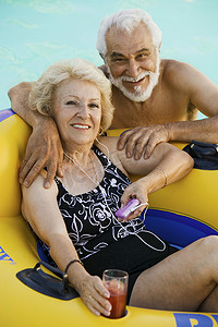 游泳池里的老夫妇女人躺在充气筏上拿着饮料听便携式音乐播放器肖像。