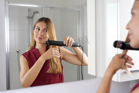 年轻拉丁女子用蒸汽直发器在浴室镜子里造型头发的特写