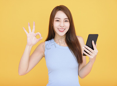 微笑的亚洲女性拿着黄色背景的手机并显示 ok 标志