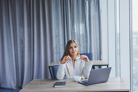 经理专注于穿着休闲装、戴着眼镜在上网本上打字的女性，同时坐在现代工作空间的桌子旁从事新项目