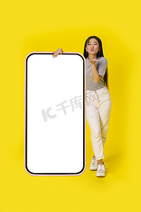 送飞吻的亚洲女孩站在巨大的智能手机旁边，智能手机上有白色屏幕，穿着休闲装，背景为黄色。