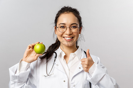 清朝皇帝画像摄影照片_年轻开朗的亚洲女医生、治疗师或糖尿病专家的画像谈论健康饮食，展示青苹果和竖起大拇指，建议吃水果，微笑鼓励照顾健康