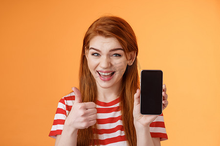 手机批准摄影照片_开朗自信的红发女性推荐智能手机应用程序，显示手机显示屏，竖起大拇指像批准标志，广泛微笑，建议良好的在线购买，移动广告