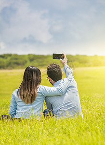 相爱的人们用智能手机在田野里自拍，微笑的相爱情侣坐在草地上自拍，年轻的相爱情侣在田野里自拍