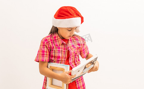 拿着照片摄影照片_戴着圣诞老人帽子的小女孩微笑着，手里拿着照片画布。