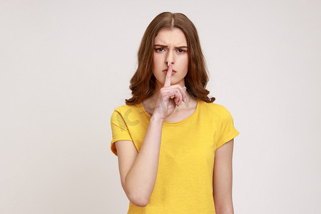 身穿黄色休闲 T 恤、严肃的棕色头发青少年女性的肖像，手指放在嘴唇上，做出安静的手势，要求保守秘密。