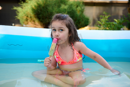 美丽的孩子，小女孩在阳光明媚的夏日在室外充气游泳池里吃冰淇淋