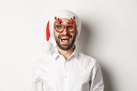 特写：戴着圣诞眼镜和圣诞帽的兴奋的大胡子男子对促销优惠、寒假广告概念、白色背景感到惊讶