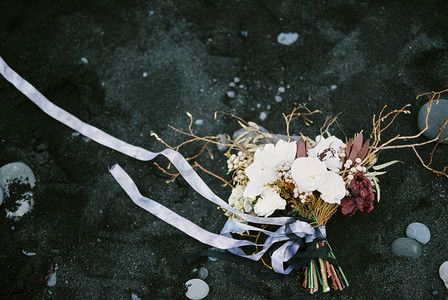 沙滩布置摄影照片_黑色的沙滩上躺着带有蓝丝带的婚礼花束。
