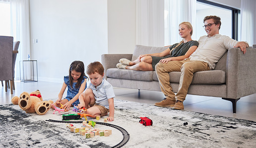 儿童玩玩具摄影照片_坐在沙发上的父母很高兴，看着孩子们在客厅地板和家里玩玩具。