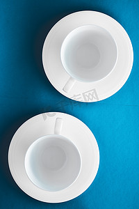 蓝色饮料摄影照片_白色餐具陶器套装，蓝色平底背景上的空杯子