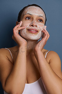 迷人的女人在脸上涂抹泡沫清洁化妆品，做按摩动作，卸妆，使用去角质美容产品使皮肤焕然一新，与灰色背景隔离