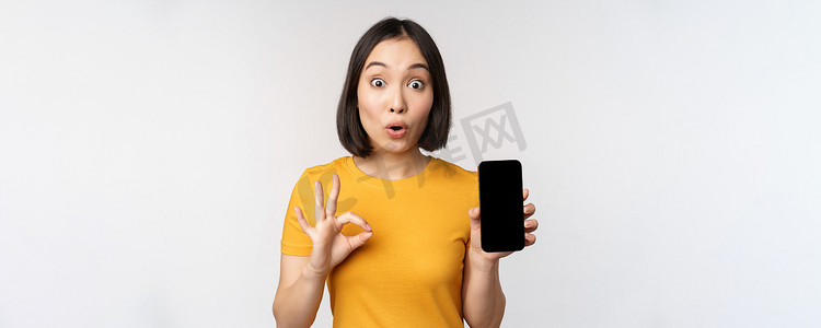 兴奋的亚洲女孩展示手机屏幕，好的标志，推荐智能手机应用程序，站在白色背景的黄色 T 恤上