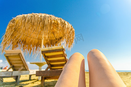 在热带沙滩上晒日光浴的女童腿，上面有模糊的稻草遮阳伞和木制太阳椅