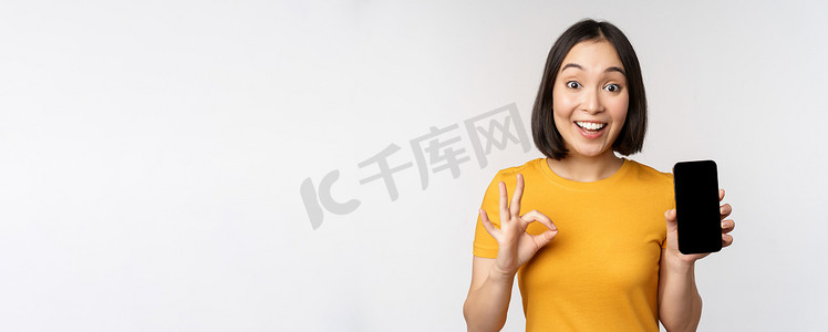 手机批准摄影照片_兴奋的亚洲女孩展示手机屏幕，好的标志，推荐智能手机应用程序，站在白色背景的黄色 T 恤上