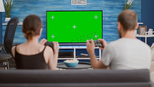 一对夫妇拿着控制器坐在沙发上，在绿屏电视上玩控制台在线游戏