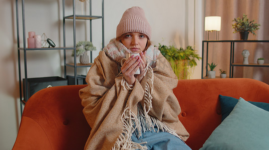 年轻生病的女人戴着格子花呢帽子，独自坐在沙发上喝着热茶，冷得瑟瑟发抖