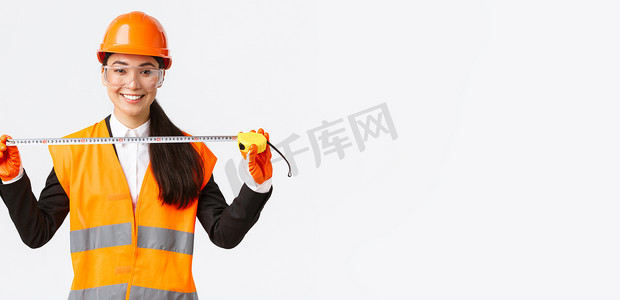 佩戴安全帽摄影照片_自信的职业亚洲女建筑师测量布局，佩戴安全帽和制服，手持卷尺，微笑高兴，对施工期间取得的成果感到满意
