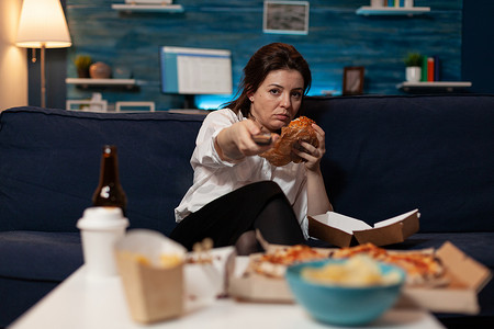 坐在沙发上吃着美味汉堡的无聊女人拿着电视上的远程切换频道
