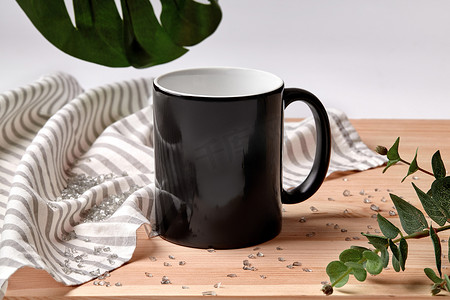 小模板摄影照片_木制桌面上的黑色瓷杯，旁边是条纹桌布、散落的水晶和白色背景下的绿色树枝。