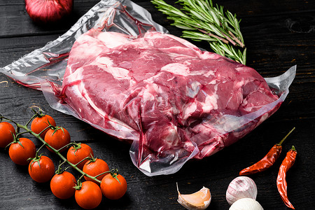 黑色木桌背景上新鲜、生鲜、真空包装的切羊肉，配料和香草