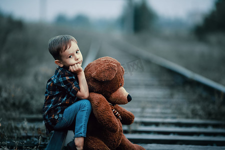 孤寂摄影照片_一个被遗弃的无家可归的孩子，一个孤儿 一个孤独的男孩抱着一个毛绒玩具，坐在铁轨上，悲伤地看着镜头