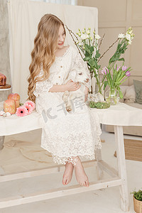 这个女孩坐在复活节餐桌旁，手里拿着蛋糕、春天的花朵和鹌鹑蛋，怀里抱着一个白色的孩子。