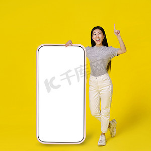 快乐的亚洲女孩靠在巨大的、巨大的智能手机上，智能手机上有白色、空白的屏幕，快乐地指着手指，穿着黄色背景的休闲装。