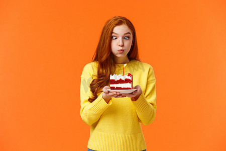 有趣而愚蠢可爱的红头发 b-day 女孩眯着眼睛做出愚蠢的表情，撅着嘴拿着生日蛋糕和吹灭蜡烛许愿，在派对庆祝中玩得开心，橙色的墙
