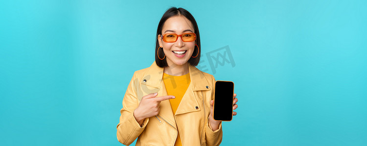 年轻时尚的亚洲女模特戴着时髦太阳镜，展示手机屏幕，在智能手机界面上应用，站在蓝色背景上