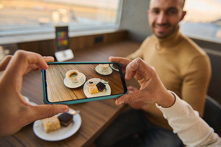 情侣旅行摄影照片_女性的手在木桌上的咖啡和蛋糕的智能手机上拍照，模糊的英俊男子坐在全景窗户前，俯瞰跑道上的飞机。