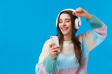 美妙音乐摄影照片_快乐的魅力和无忧无虑的微笑欧洲女性在作为圣诞礼物收到的新耳机中享受美妙的音乐质量声音，举起手拿着智能手机，蓝色背景