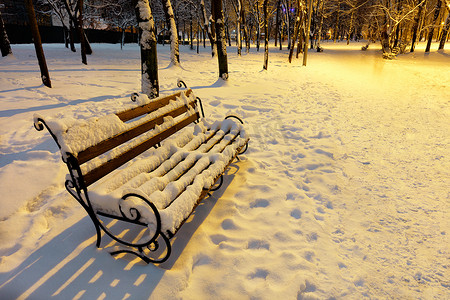 冬季城市夜间公园的木凳上覆盖着雪，并被路灯的暖光照亮。