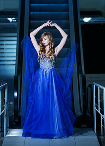 美女后面摄影照片_身着飘逸的蓝色晚礼服、背景是移动楼梯的金发美女