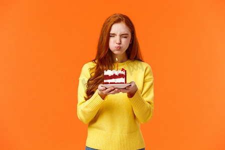 橙色生日摄影照片_可爱又傻，可爱的红发生日女孩闭上眼睛，拿着生日蛋糕，撅着嘴吹蜡烛许愿，希望梦想成真，闭上眼睛，和朋友一起庆祝，橙色背景
