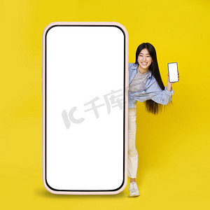 巨大手机摄影照片_可爱的亚洲年轻女孩兴奋地偷看巨大的垂直手机，手里拿着较小的手机，白色空白屏幕与黄色背景隔离。