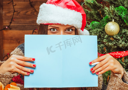 戴着圣诞老人帽的白人模特女性用一张蓝色的纸遮住脸的下半部分，手里拿着笔。