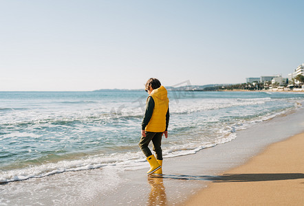 穿着黄色橡胶靴的男孩在海滩玩水和沙子。
