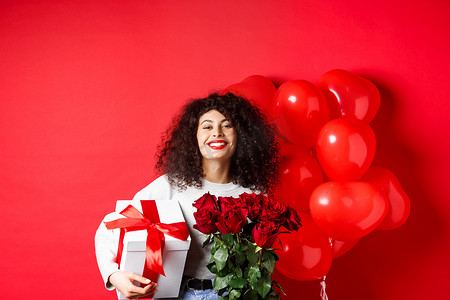 微笑的快乐女人拿着盒子，里面装着男朋友的礼物和红玫瑰，庆祝情人节，站在浪漫的心形气球旁，站在工作室背景上